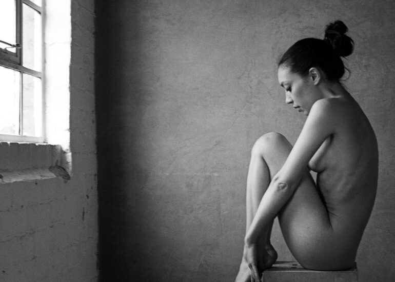 Fine art nude photograph of a woman posed beside a Victorian window, using natural light to sculpt form and shadow.