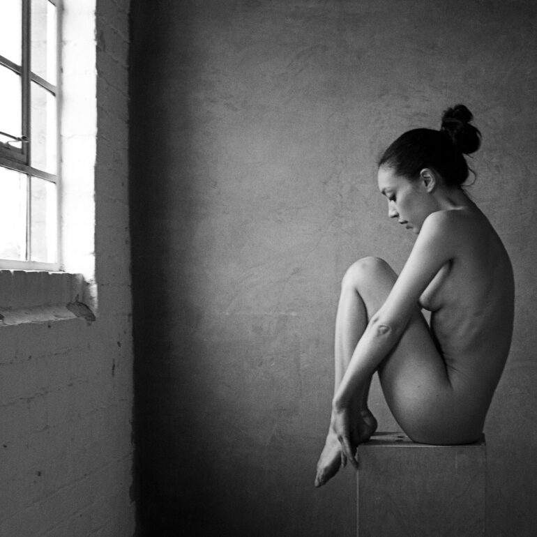 Fine art nude photograph of a woman posed beside a Victorian window, using natural light to sculpt form and shadow.