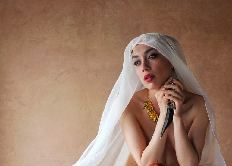 Fine art nude photograph of a veiled woman resting at a table with an apple and dagger, expressing tension and reflection.