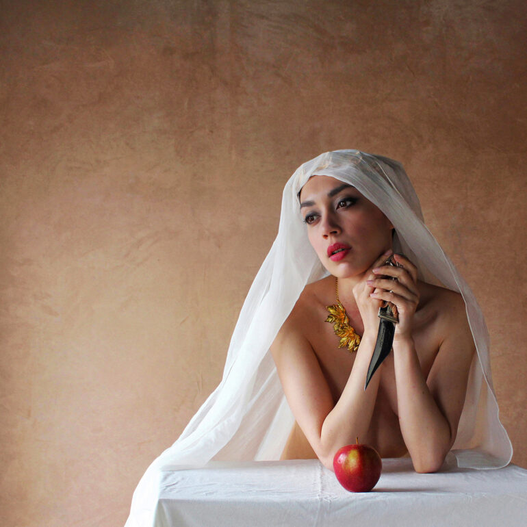 Fine art nude photograph of a veiled woman resting at a table with an apple and dagger, expressing tension and reflection.