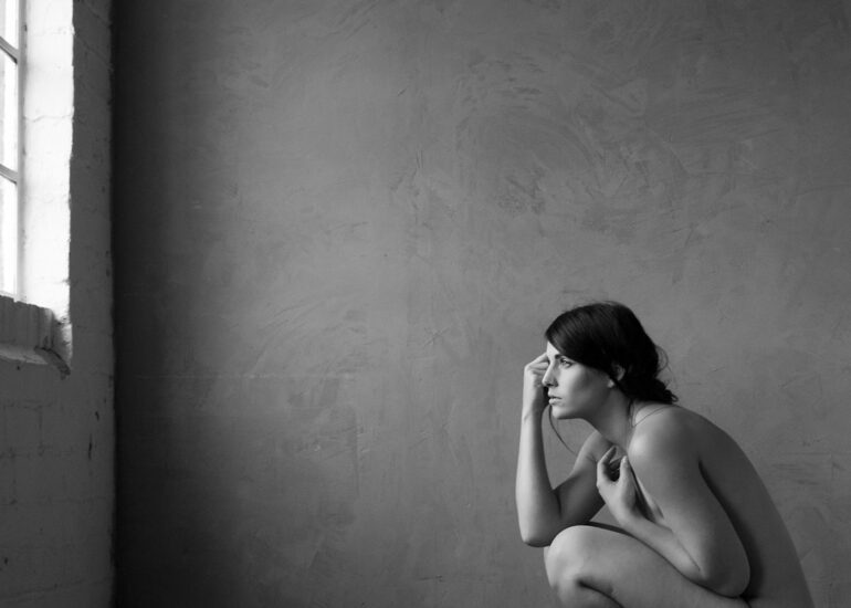 Fine art nude photograph of a crouched woman gazing toward a window, expressing introspection, vulnerability, and quiet emotion.
