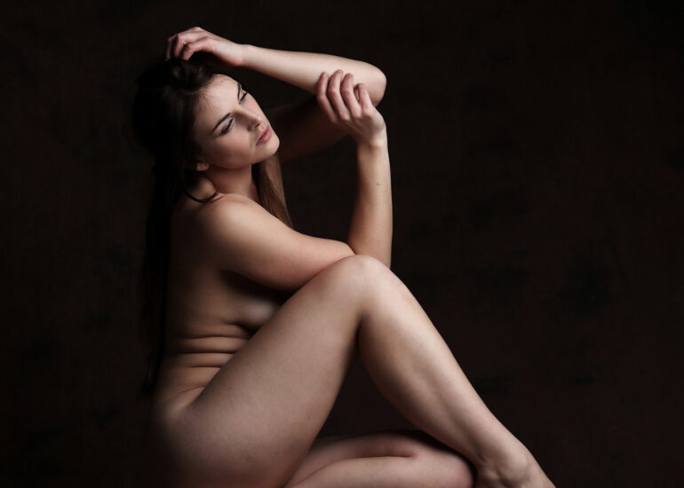 Fine art nude photograph of a woman seated alone against a dark backdrop, focusing on form, stillness, and emotional quiet.