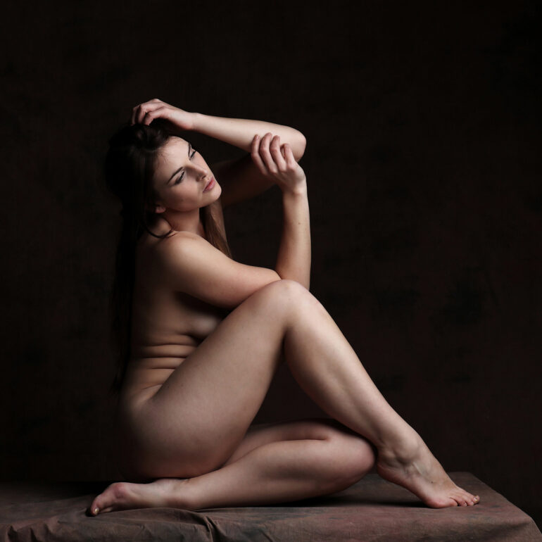 Fine art nude photograph of a woman seated alone against a dark backdrop, focusing on form, stillness, and emotional quiet.