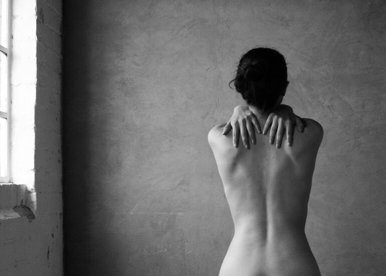 Minimalist black and white fine art nude showing a woman embracing herself in soft natural window light.