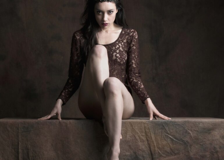 Fine art nude portrait of a regal woman seated on a plinth, wearing a crown and lace garment, expressing power, dominance, and dark feminine authority in a classical studio setting.