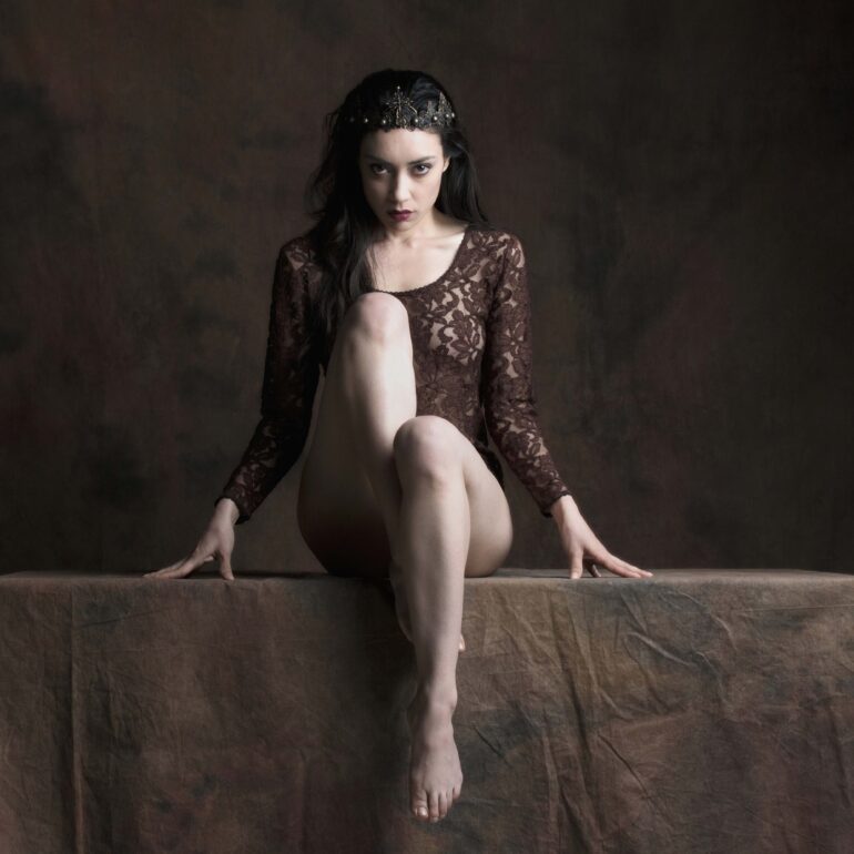 Fine art nude portrait of a regal woman seated on a plinth, wearing a crown and lace garment, expressing power, dominance, and dark feminine authority in a classical studio setting.