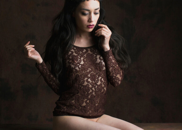 Fine art portrait of a dark-haired woman wearing a delicate crown and lace garment, seated in a classical studio setting, expressing quiet power, elegance, and feminine introspection