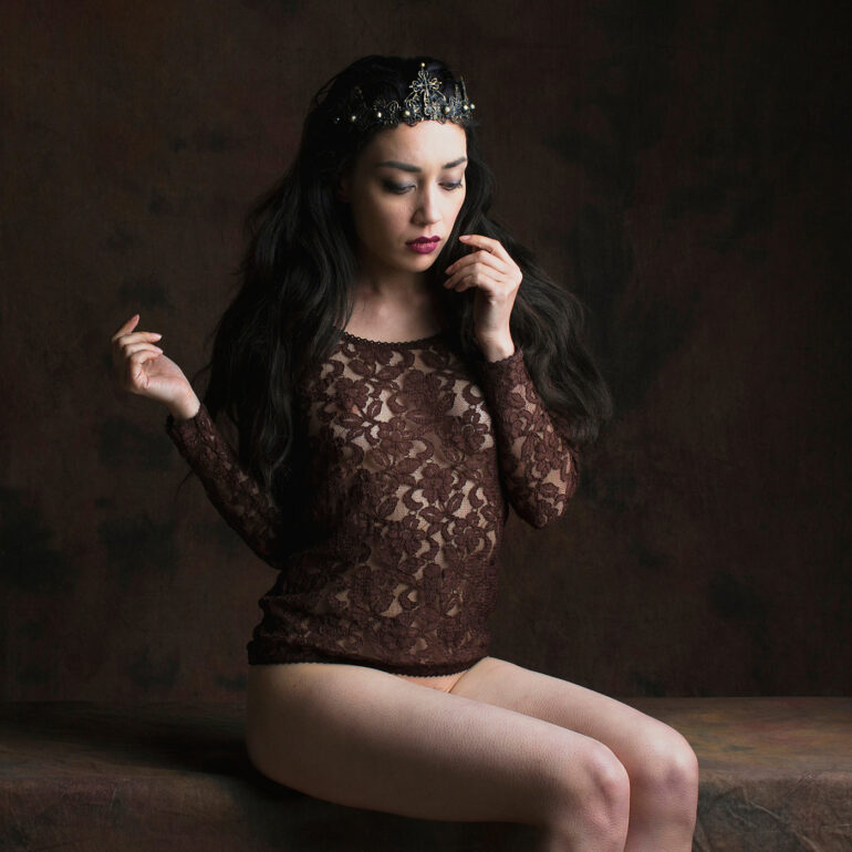 Fine art portrait of a dark-haired woman wearing a delicate crown and lace garment, seated in a classical studio setting, expressing quiet power, elegance, and feminine introspection