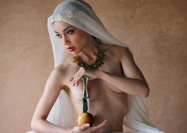 Fine art nude photograph of a veiled woman holding an Arabian dagger, illuminated by soft natural light against a textured backdrop, exploring mystery, ritual, and classical symbolism.