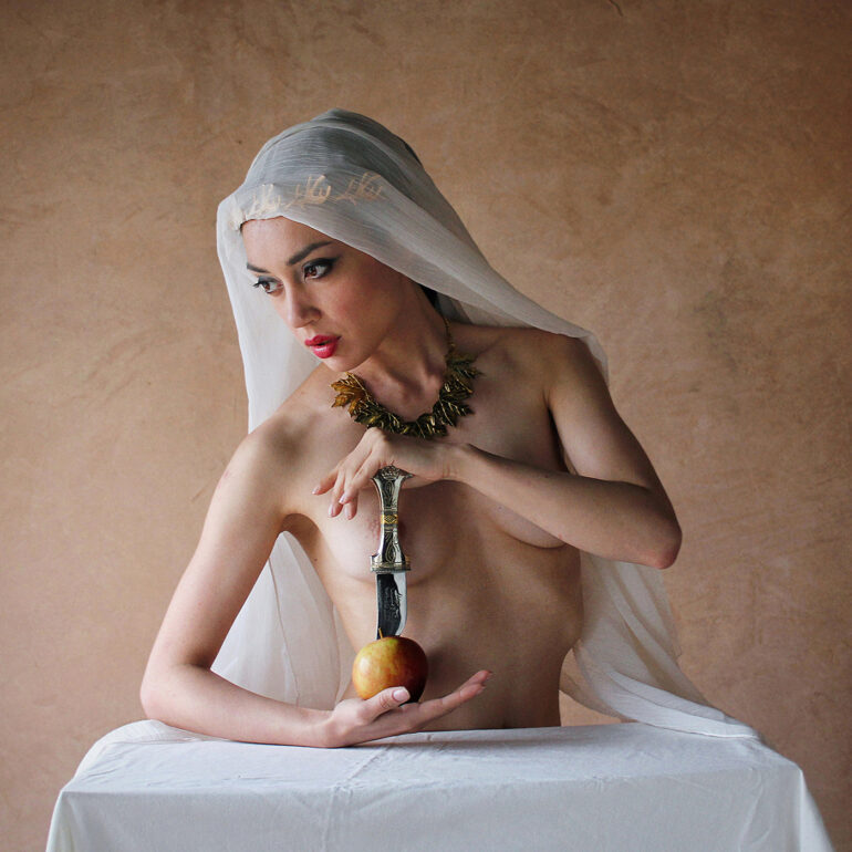 Fine art nude photograph of a veiled woman holding an Arabian dagger, illuminated by soft natural light against a textured backdrop, exploring mystery, ritual, and classical symbolism.
