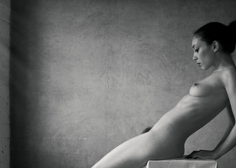 Black and white fine art nude photograph of a seated woman illuminated by soft window light, emphasising form, intimacy, and stillness.