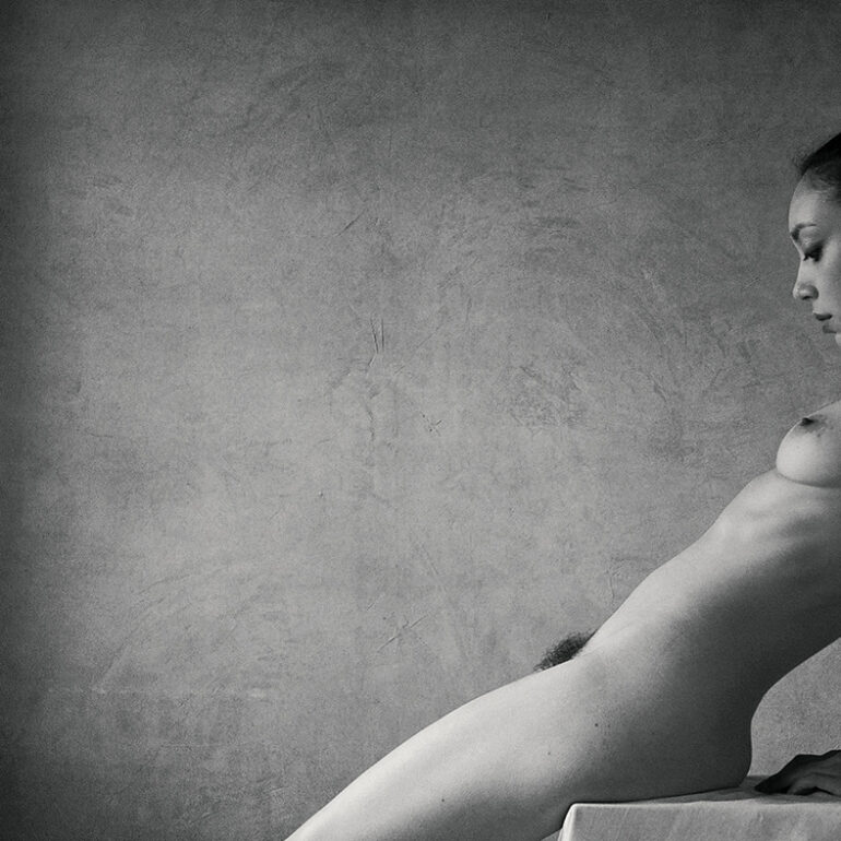 Black and white fine art nude photograph of a seated woman illuminated by soft window light, emphasising form, intimacy, and stillness.