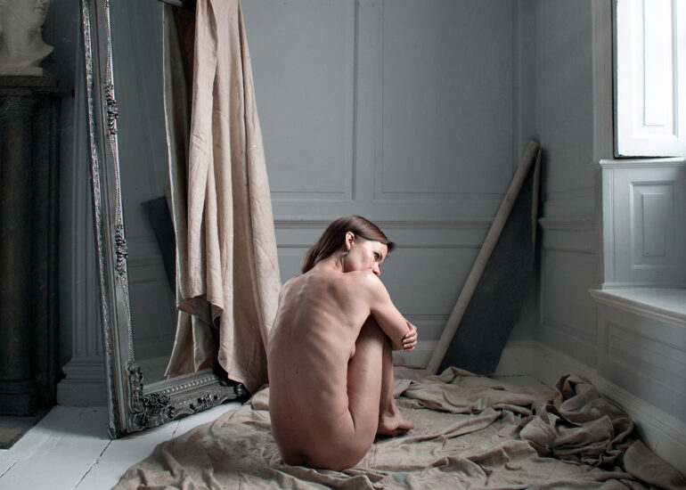Award-winning fine art nude photograph of a naked woman sitting alone on draped fabric in a classical studio interior, illuminated by soft natural window light.