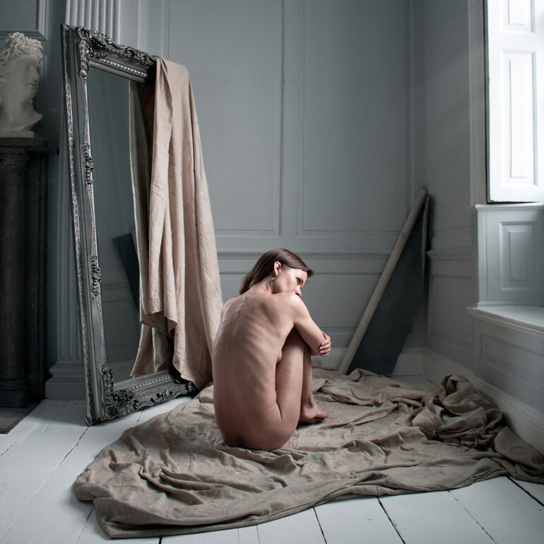 Award-winning fine art nude photograph of a naked woman sitting alone on draped fabric in a classical studio interior, illuminated by soft natural window light.